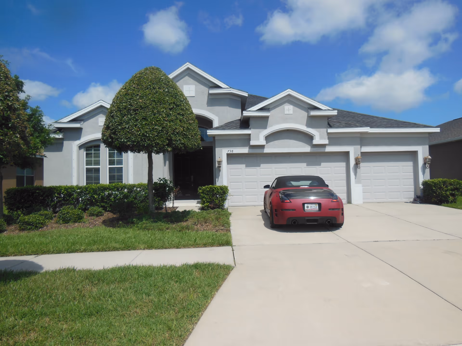 Front exterior view of a single-story house with a neatly trimmed tree and bushes in the front yard, a three-car garage, and a red convertible car parked in the driveway under a blue sky with scattered clouds.