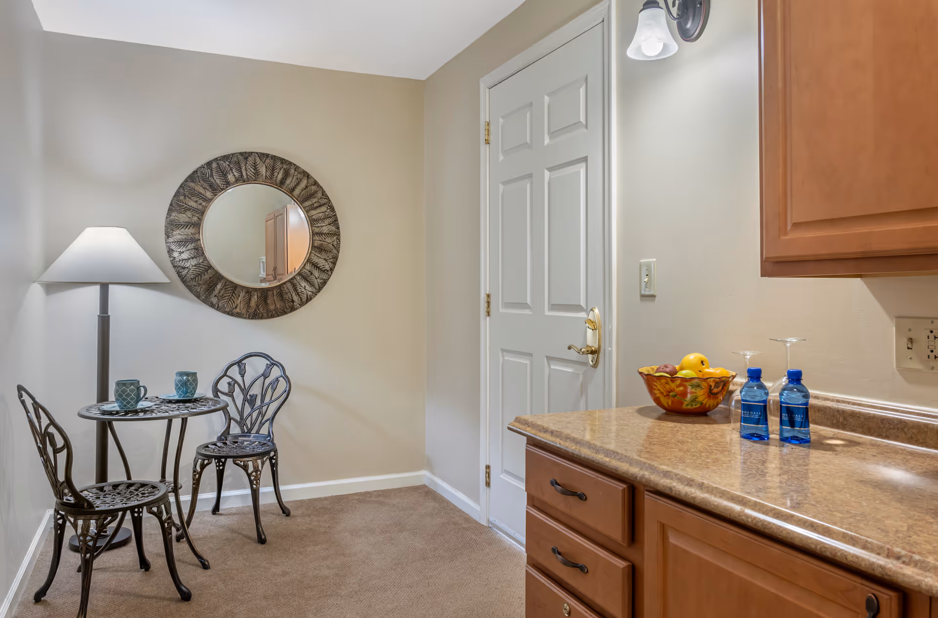 Small dining area with a round metal table and two matching chairs, a floor lamp beside the table, a decorative round mirror on the wall, and a kitchen counter with a bowl of fruit and two bottles of water.