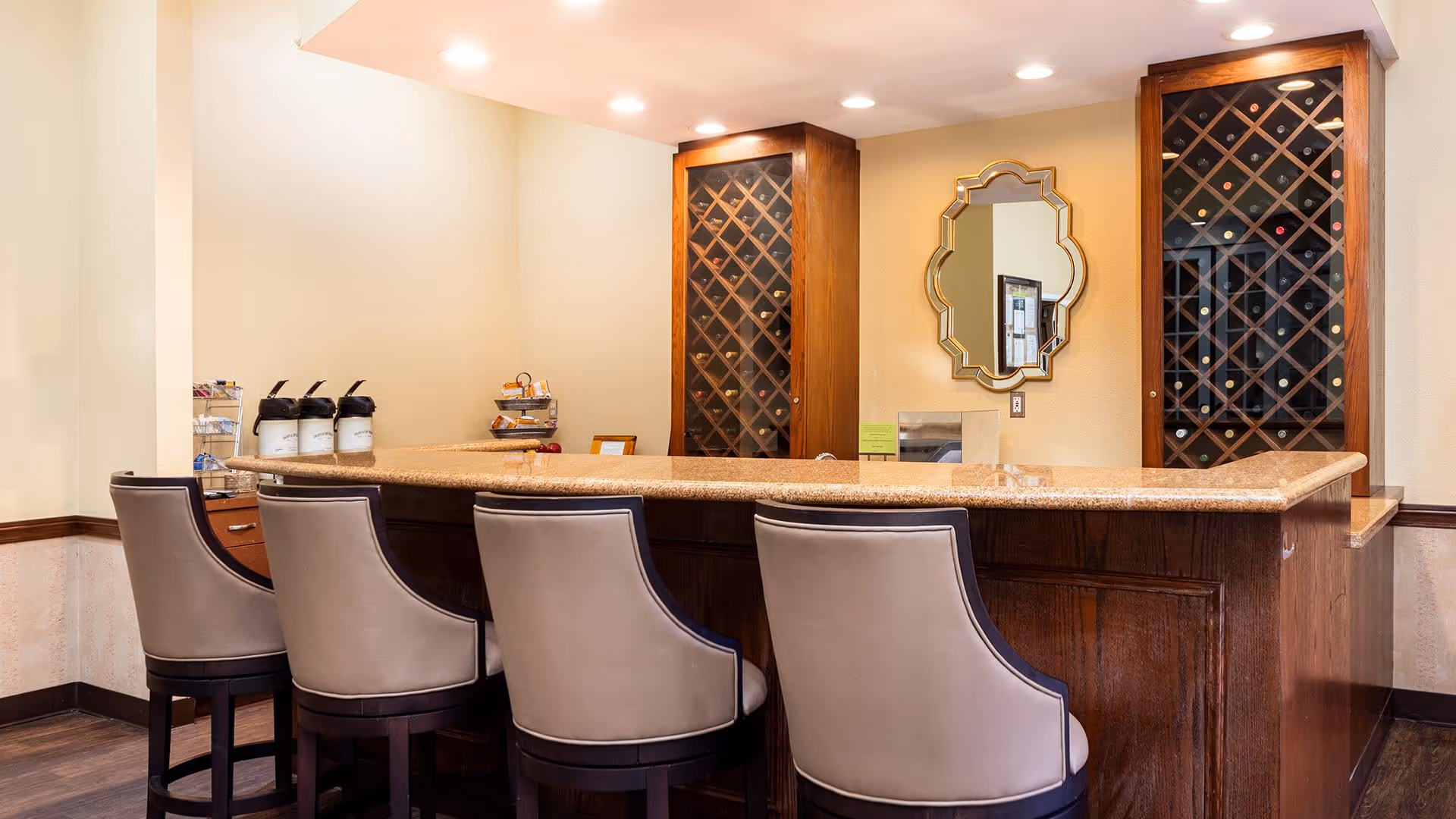 Interior view of a bar area with four cushioned bar stools in front of a wooden counter with a granite top. Behind the counter are two wooden wine racks filled with bottles and a decorative mirror on the wall. The area is well-lit with recessed ceiling lights.