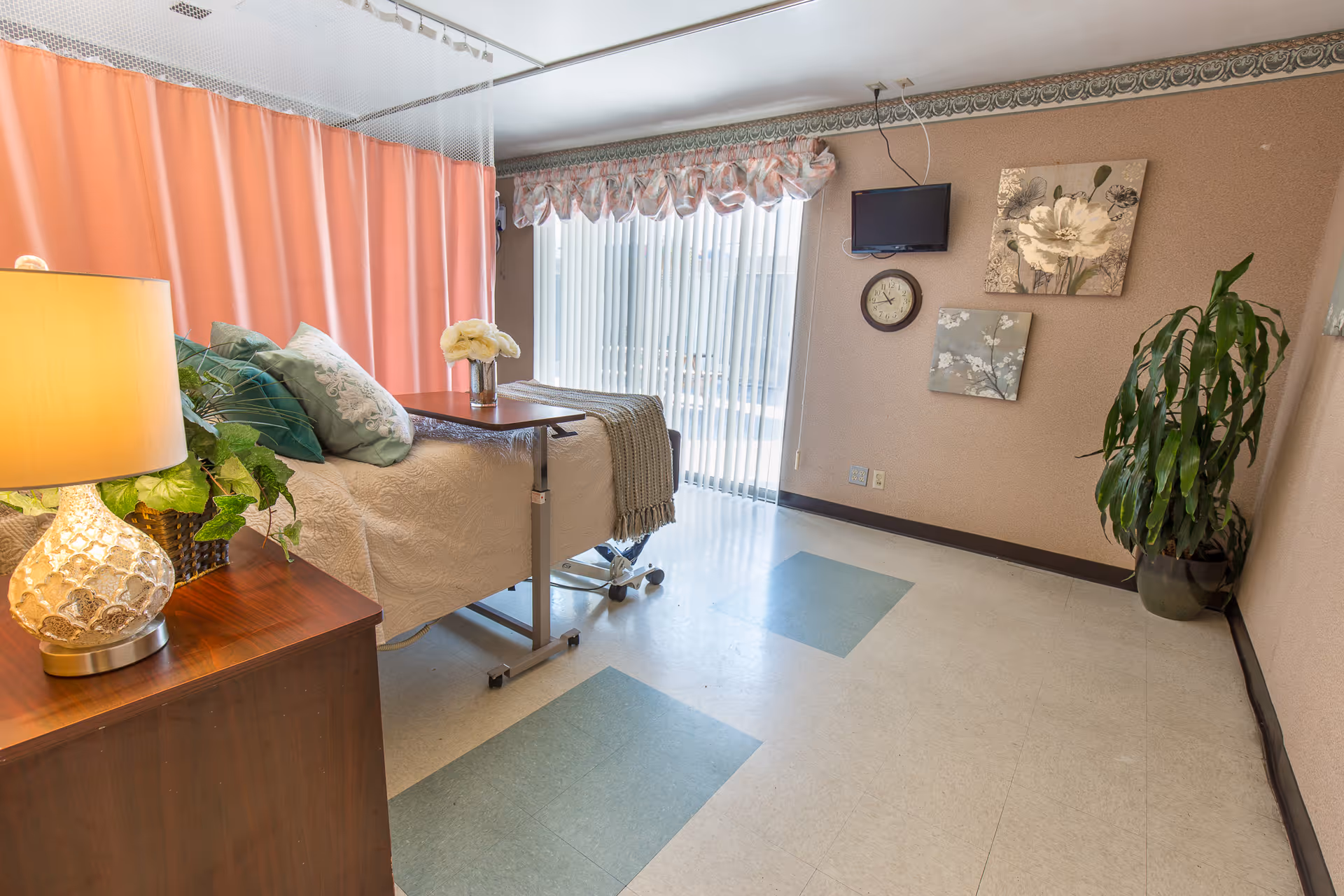 A bright and clean patient room with a hospital bed covered in a beige quilt and green pillows, a wooden bedside table with a decorative lamp and plant, a pink privacy curtain, a wall-mounted TV, a clock, floral wall art, a large potted plant, and vertical blinds covering a window or glass door.