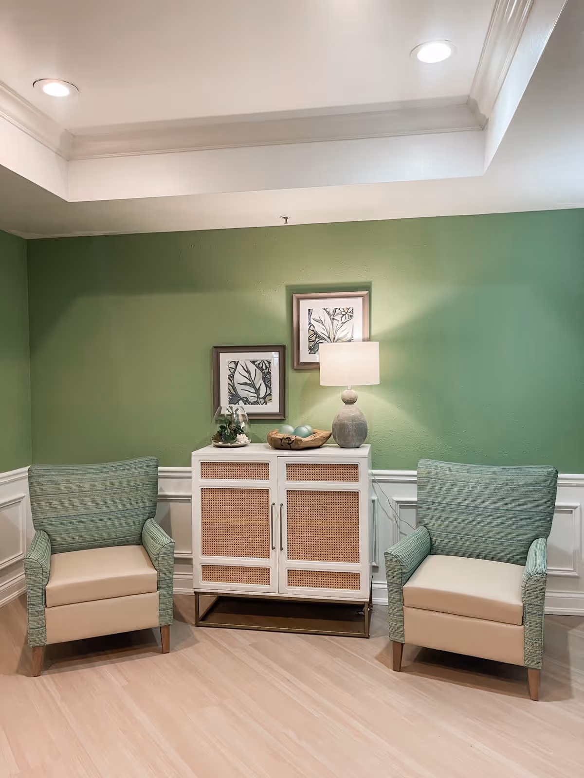 A cozy corner with two green and beige armchairs flanking a white cabinet with wicker panel doors. On top of the cabinet is a table lamp, a decorative bowl with blue spheres, and a small plant. Two framed botanical prints hang on the green wall behind the cabinet. The floor is light wood, and the ceiling has recessed lighting.