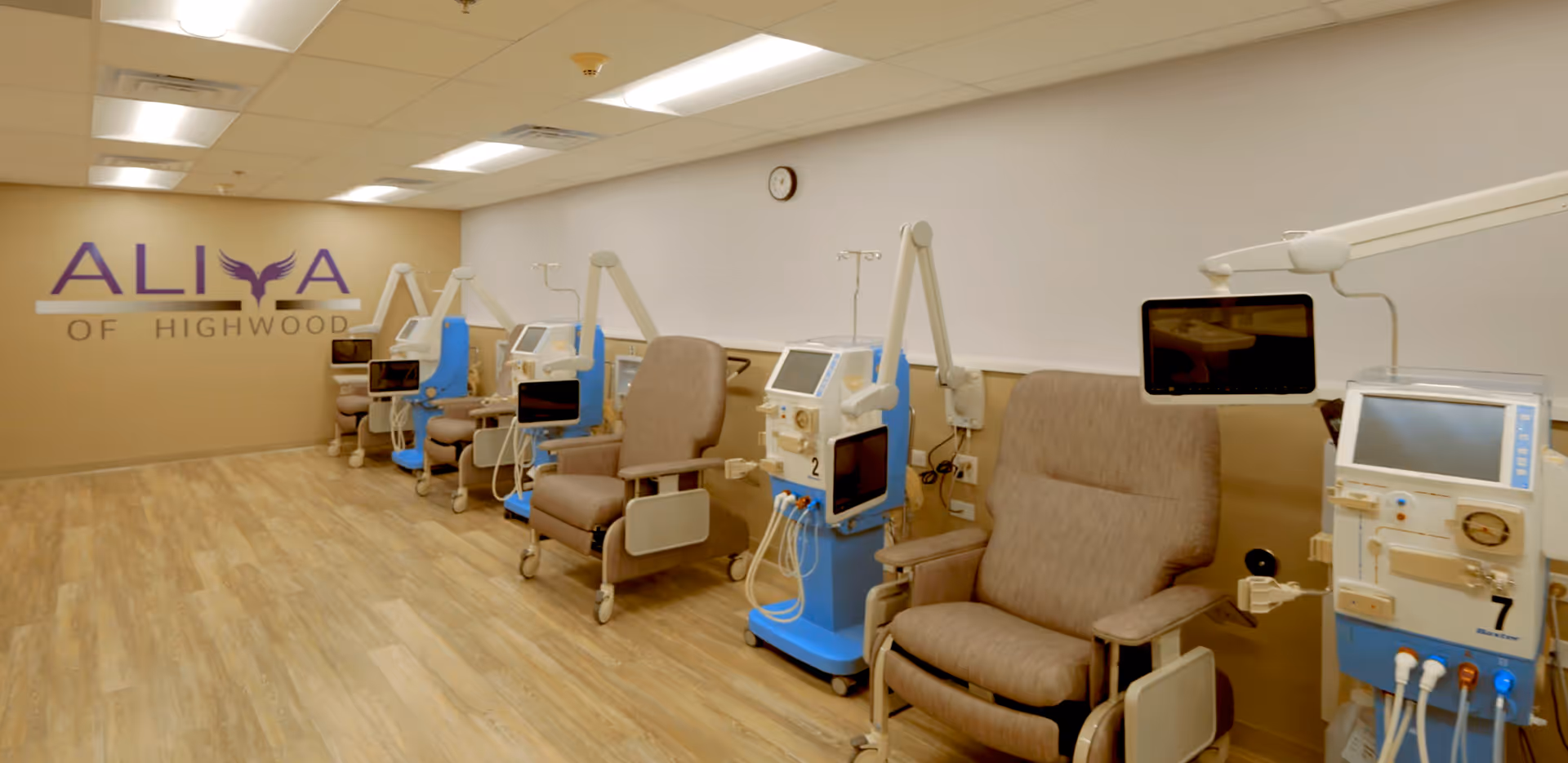 A medical treatment room at ALIYA of Highwood featuring a row of comfortable recliner chairs each paired with dialysis machines and attached monitors. The room has wood-patterned flooring, beige walls, and a clock on the wall.