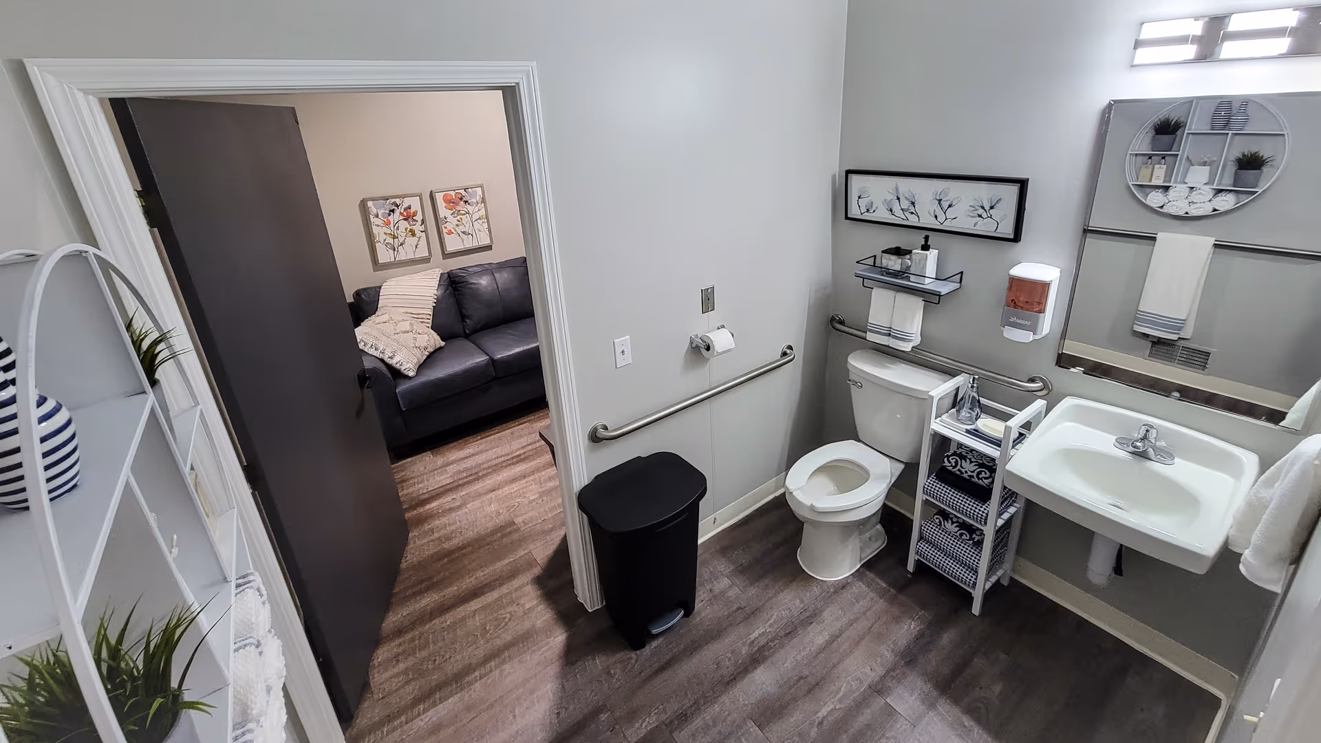 A clean and accessible bathroom with a white toilet, sink, and a large mirror above the sink. There are handrails next to the toilet and a small shelving unit with towels and toiletries. The bathroom has wood-look flooring and light gray walls. The door is open, showing a glimpse of a living room with a dark couch and floral wall art. A white circular shelf with plants and towels is mounted on the wall above the toilet.