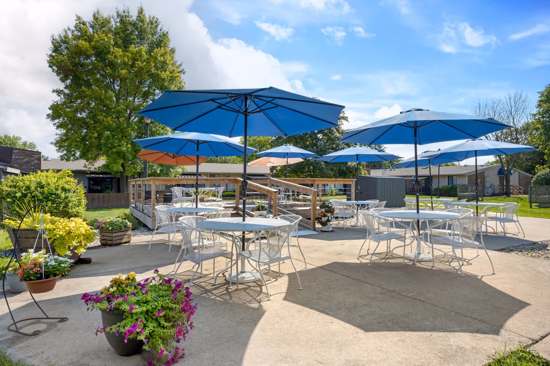 Outdoor patio with white metal tables and chairs under blue umbrellas and potted flowers.