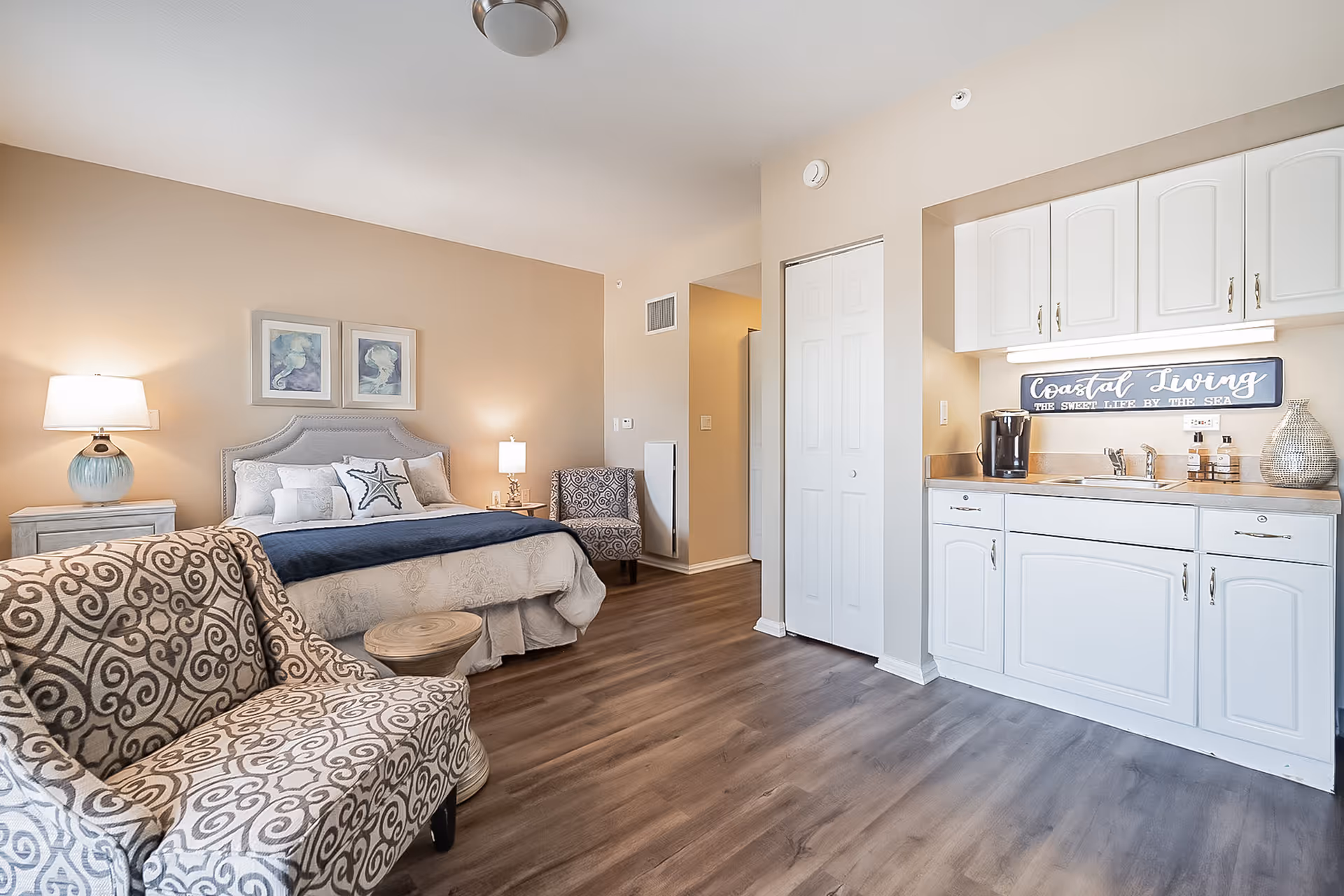A cozy studio apartment with a bed featuring white and blue bedding, two framed artworks above the headboard, a patterned armchair with a small round wooden table in front, and a kitchenette with white cabinets, a sink, a coffee maker, and a decorative sign that reads 'Coastal Living The Sweet Life By The Sea'. The room has wood flooring and beige walls with soft lighting from two lamps.