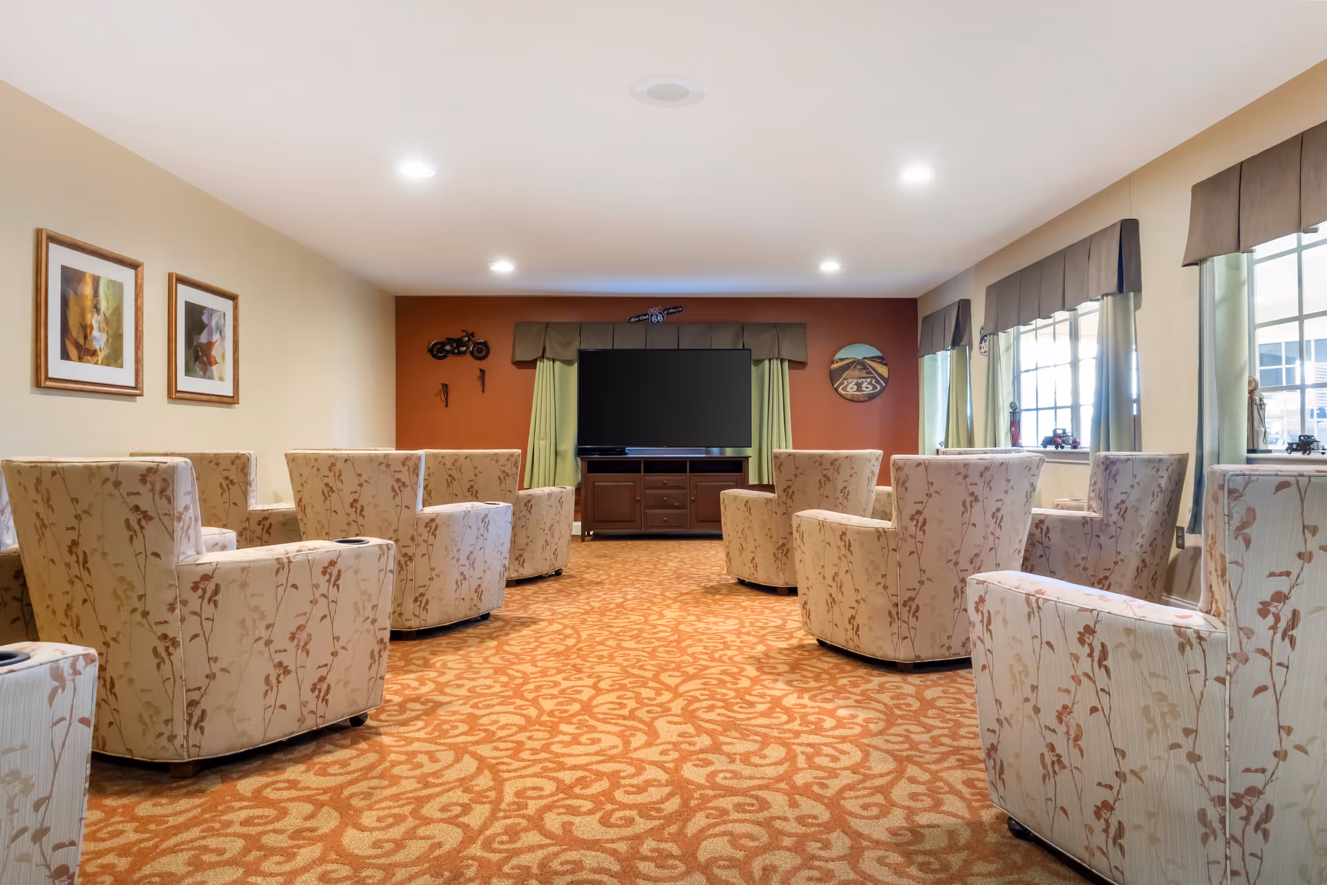 Carpeted lounge with patterned swivel chairs arranged facing a large TV and windows in a senior living facility common room.