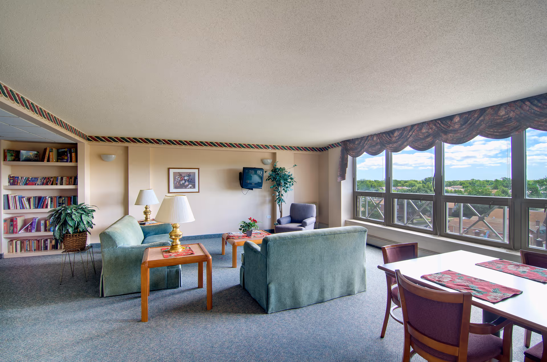 A spacious living room area with large windows offering a view of trees and rooftops outside. The room features two green upholstered sofas, a wooden coffee table, a side table with a lamp, a single armchair, a wall-mounted TV, a bookshelf filled with books, and a dining table with chairs and placemats near the windows. The room is carpeted and decorated with a patterned valance above the windows.