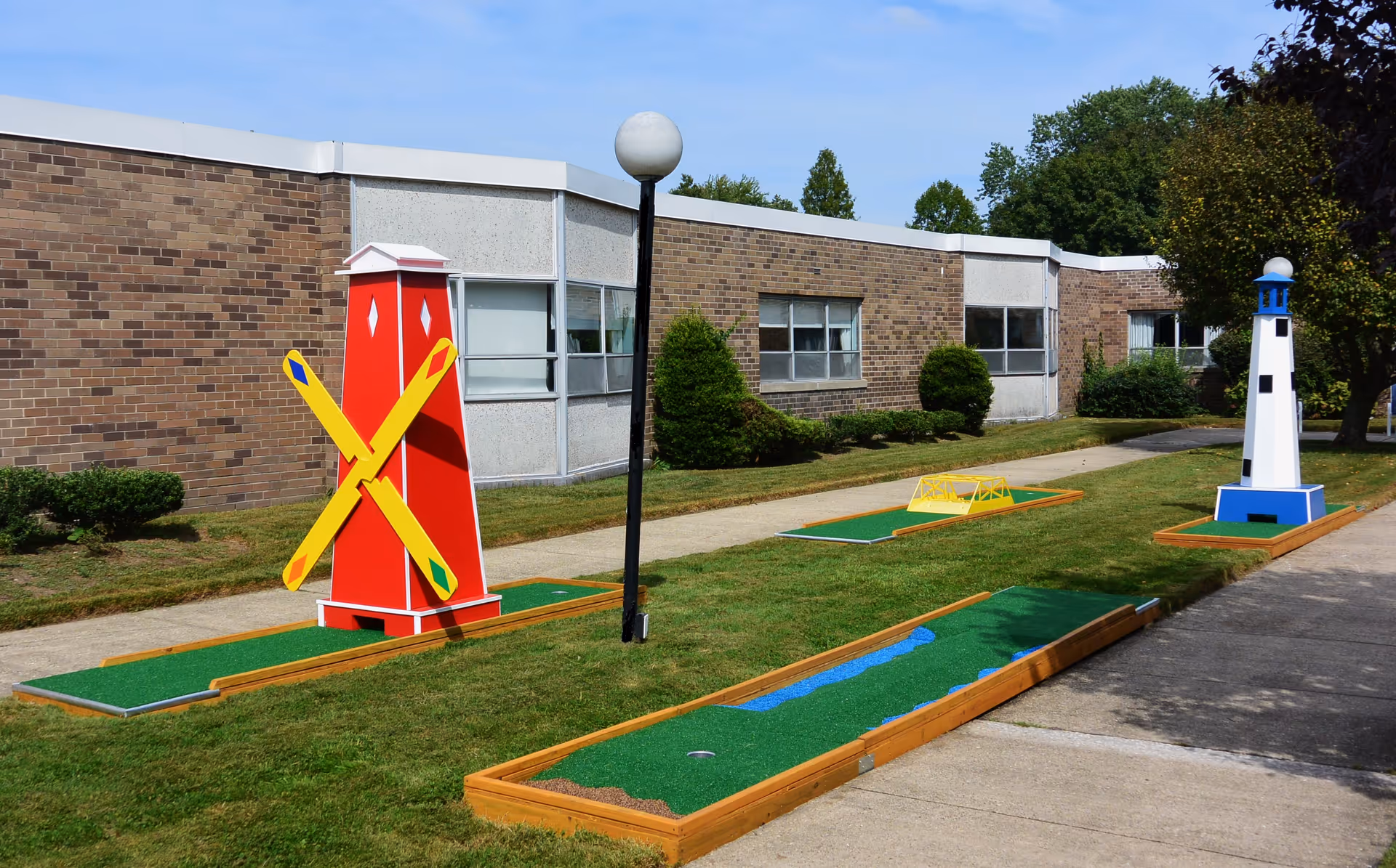 Outdoor miniature golf course with colorful obstacles including a red windmill with yellow blades and a blue and white lighthouse, situated on a grassy area next to a brick building with windows and a sidewalk.