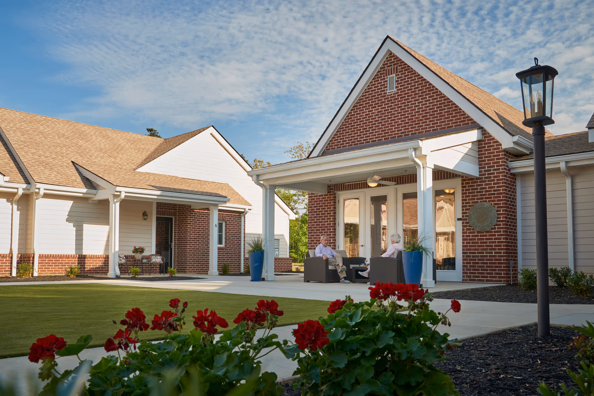 Two elderly people sitting and talking on outdoor chairs under a covered porch of a brick and white siding building with a well-maintained lawn and red flowers in the foreground.