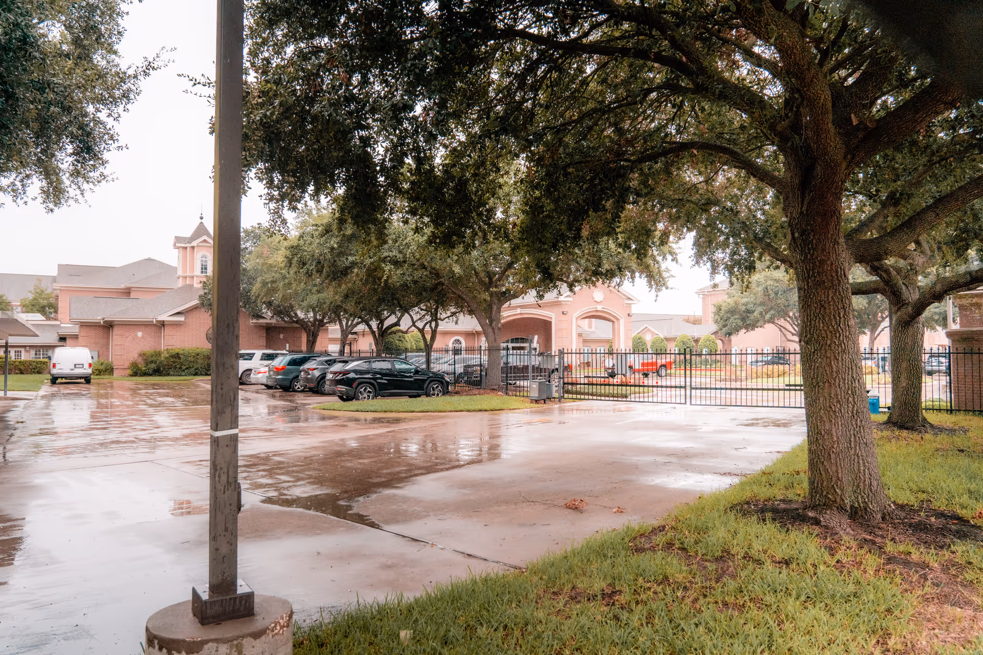 Gated entrance and wet parking lot in front of a brick senior living building, with parked cars and large oak trees.