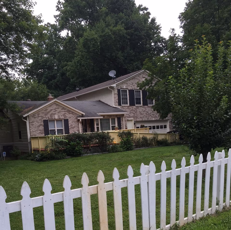 Brick house with a white picket fence, deck, and green lawn.