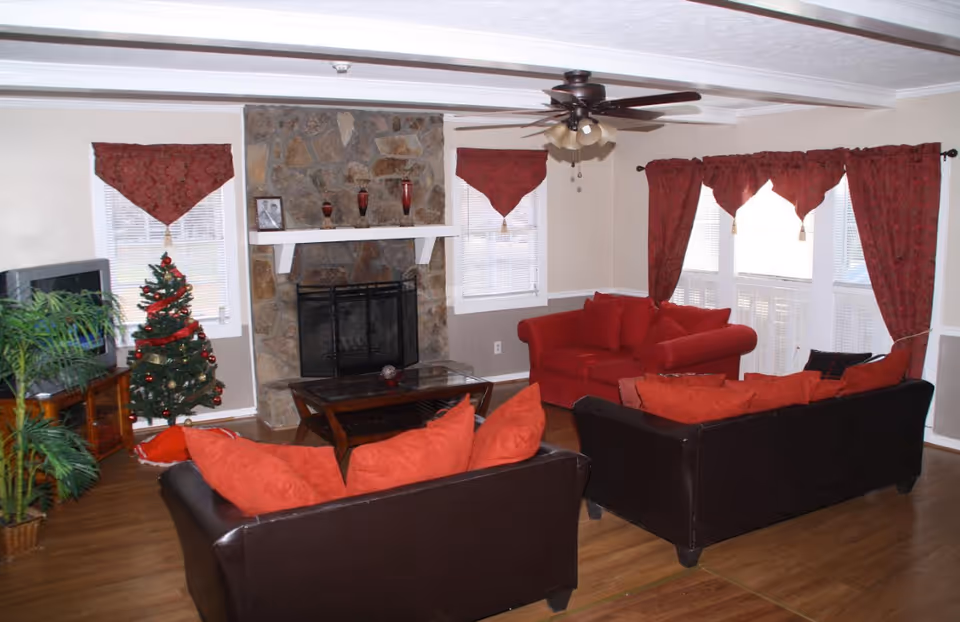 A cozy living room with two dark brown sofas and one red sofa, all adorned with red cushions. There is a stone fireplace in the center of the room with decorative items on the mantel. The room has wooden flooring, a ceiling fan with lights, and windows with red curtains and valances. A small decorated Christmas tree is placed near a TV on a wooden stand, and there are some green plants in the corner.