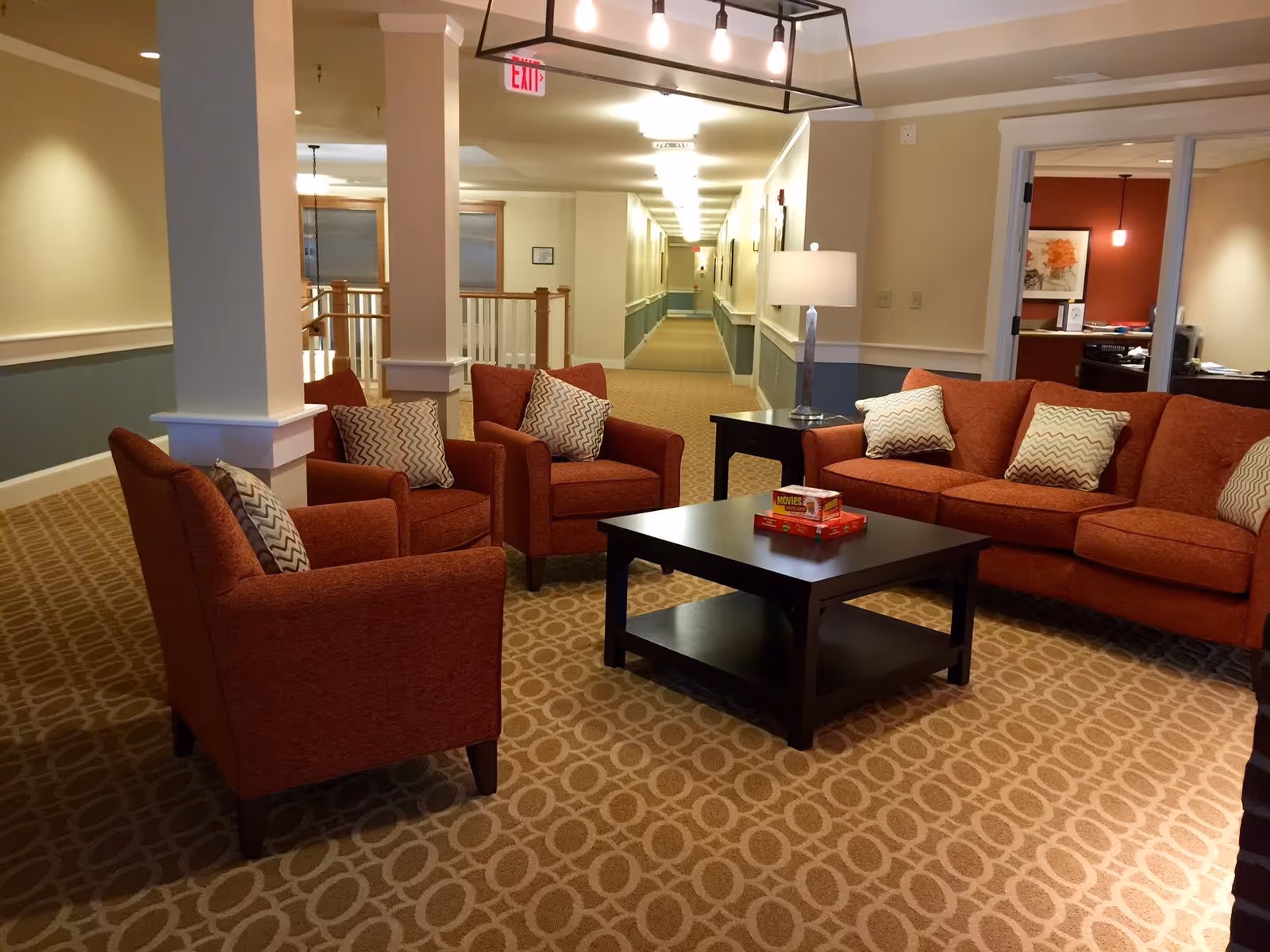 A cozy seating area in a senior living facility with a red sofa, three matching armchairs, and a dark wooden coffee table with board games on it. The room has patterned carpet, a table lamp, and a long hallway in the background.