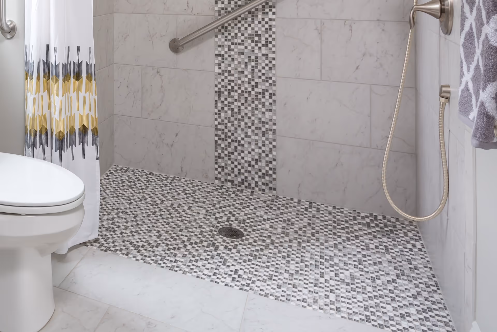 A bathroom shower area with a tiled floor and wall featuring a vertical strip of small mosaic tiles. There is a handheld showerhead attached to the wall, a grab bar for support, a white toilet partially visible on the left, and a white shower curtain with a yellow and gray pattern.