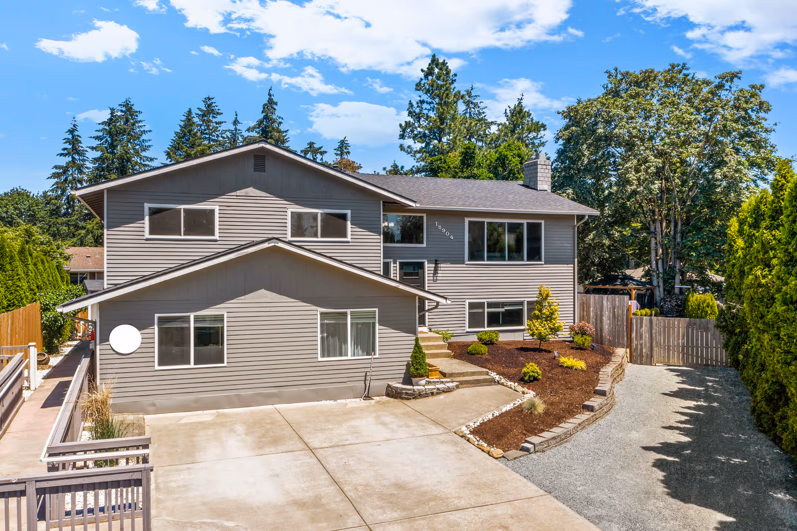 Exterior view of a two-story gray house with multiple windows, a concrete driveway, and a landscaped front yard with small shrubs and trees under a blue sky with scattered clouds.