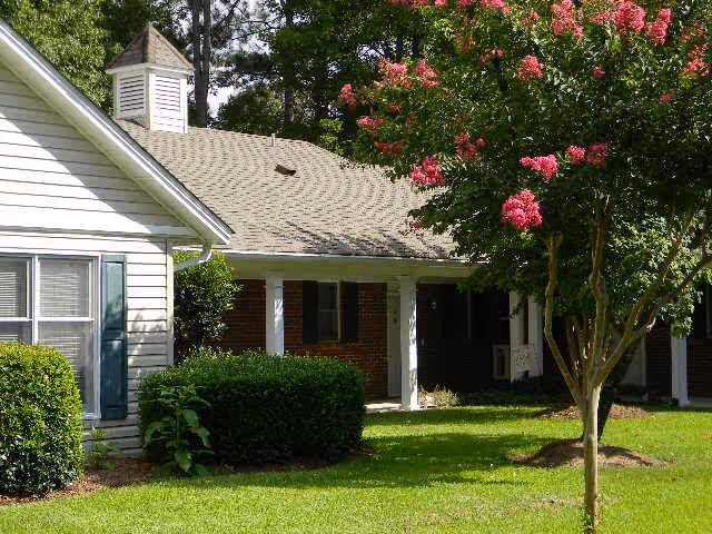 Exterior view of a residential building with white siding and brick walls, surrounded by green bushes and a tree with pink flowers on a sunny day.