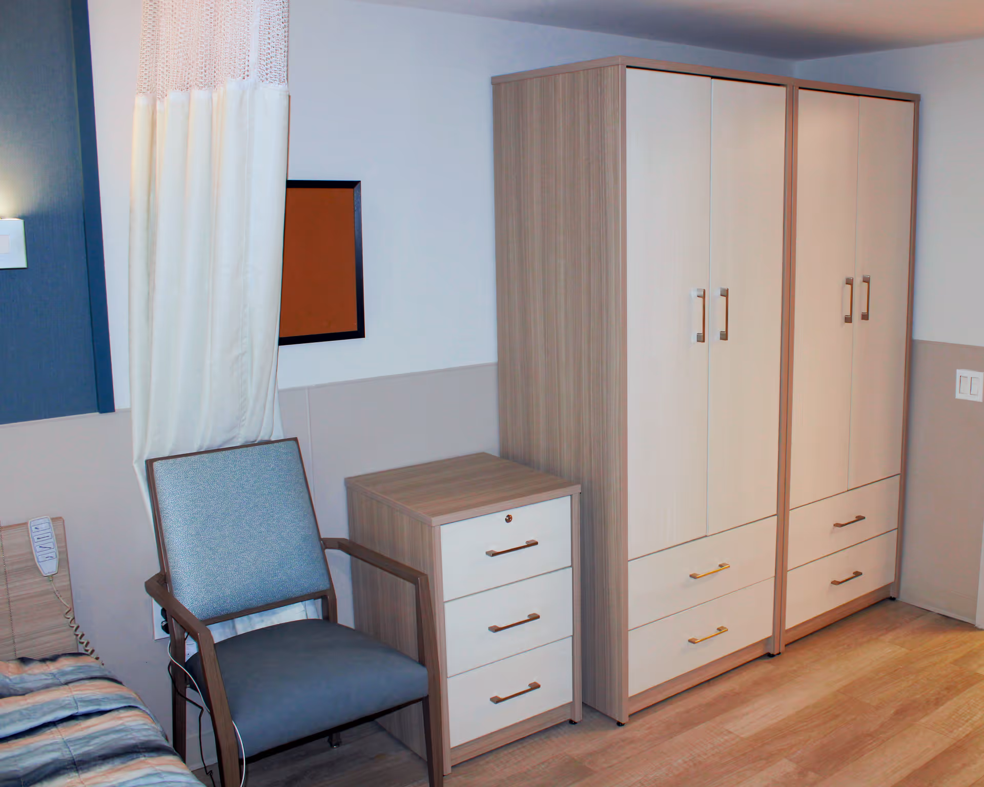 A corner of a bedroom in a senior living facility showing a wooden wardrobe with multiple doors and drawers, a matching nightstand with three drawers, and a blue upholstered chair with wooden armrests. Part of a bed with a striped blanket and a privacy curtain are also visible.