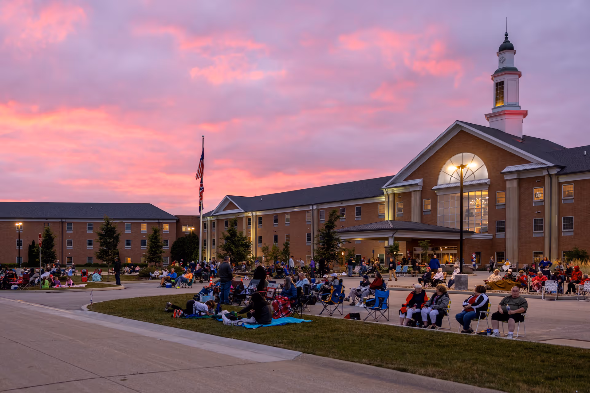 A large group of people gathered outside the Western Reserve Masonic Community building during sunset, sitting on chairs and blankets on the grass and pavement, with a colorful pink and purple sky in the background.