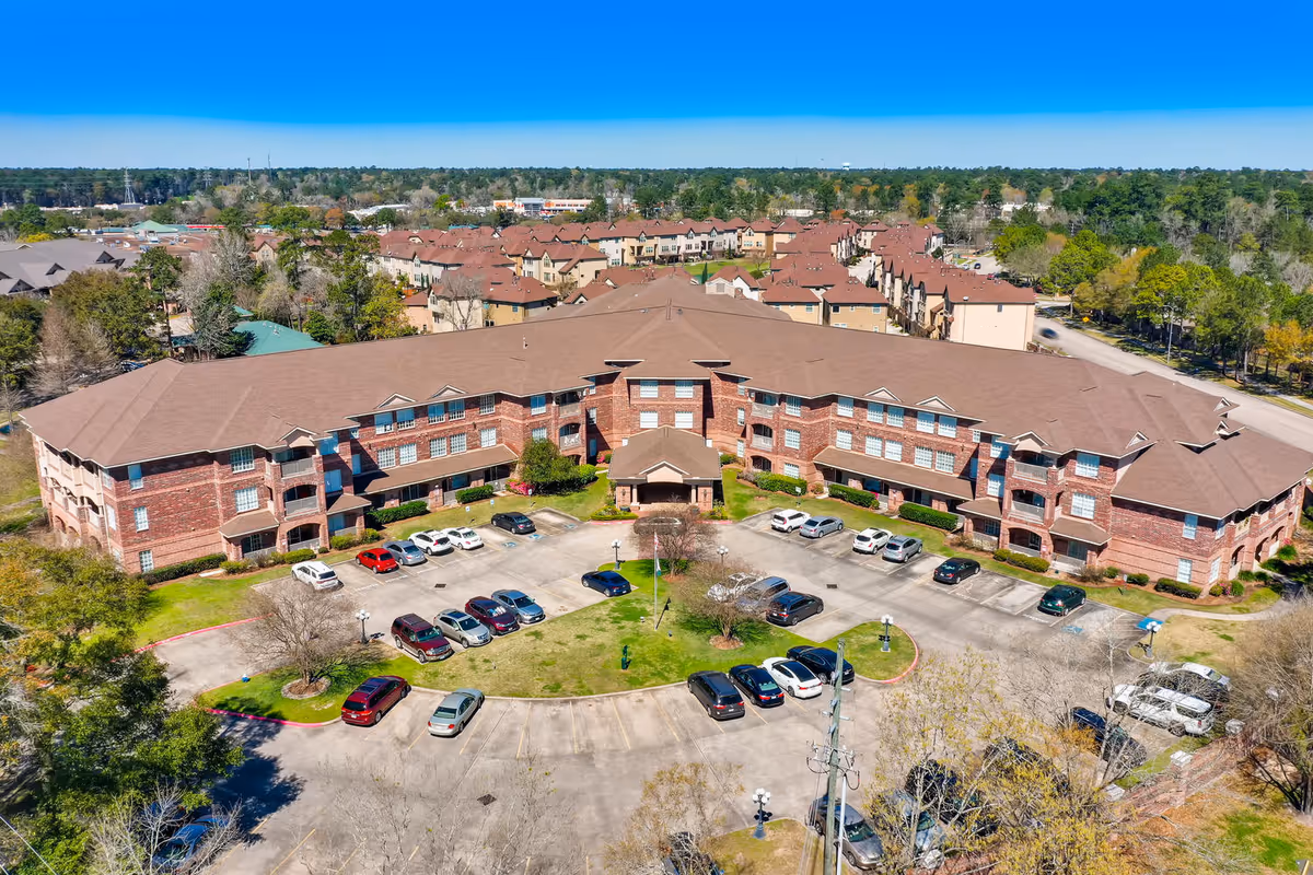 Aerial view of a three-story brick senior living building with a semicircular driveway and parked cars, surrounded by trees and neighboring residences.