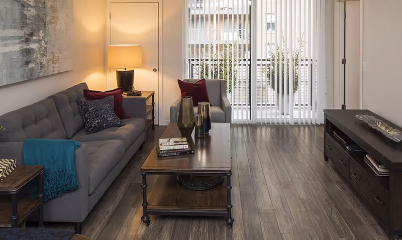Well-lit living room with a gray tufted sofa, armchair, wooden coffee table and TV console in front of vertical blinds covering a sliding glass door.