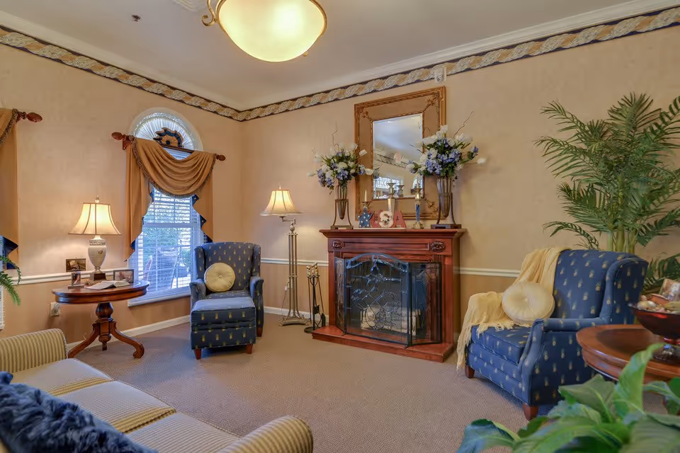 A cozy living room with beige walls and carpet, featuring a wooden fireplace with a decorative screen and a large mirror above it. Two blue armchairs with patterned upholstery and round cushions flank the fireplace. There are two side tables with lamps, a beige sofa, and several plants adding greenery to the space. The window has an arched top with draped curtains.
