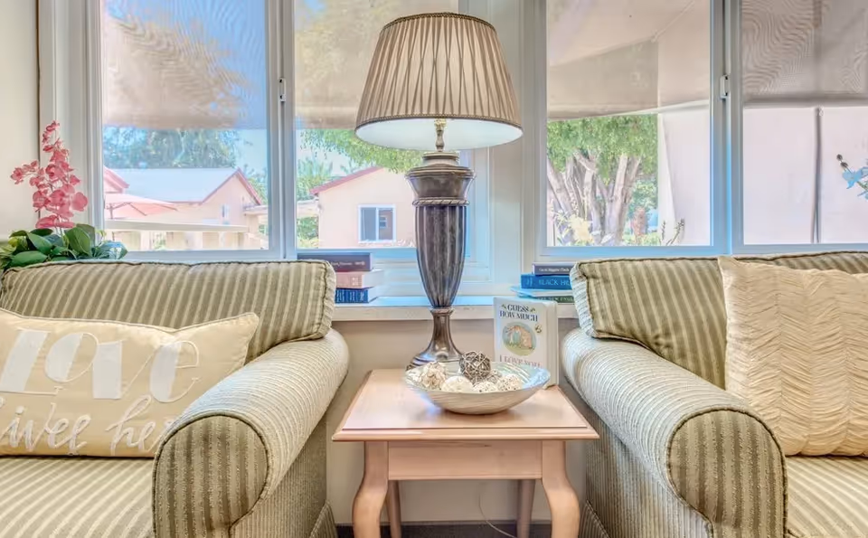 A cozy living room area with two striped armchairs facing each other, separated by a wooden side table holding a decorative bowl with woven balls and a tall table lamp. Behind the table is a window with a view of trees and houses outside. There are books and a small plant on the window sill.