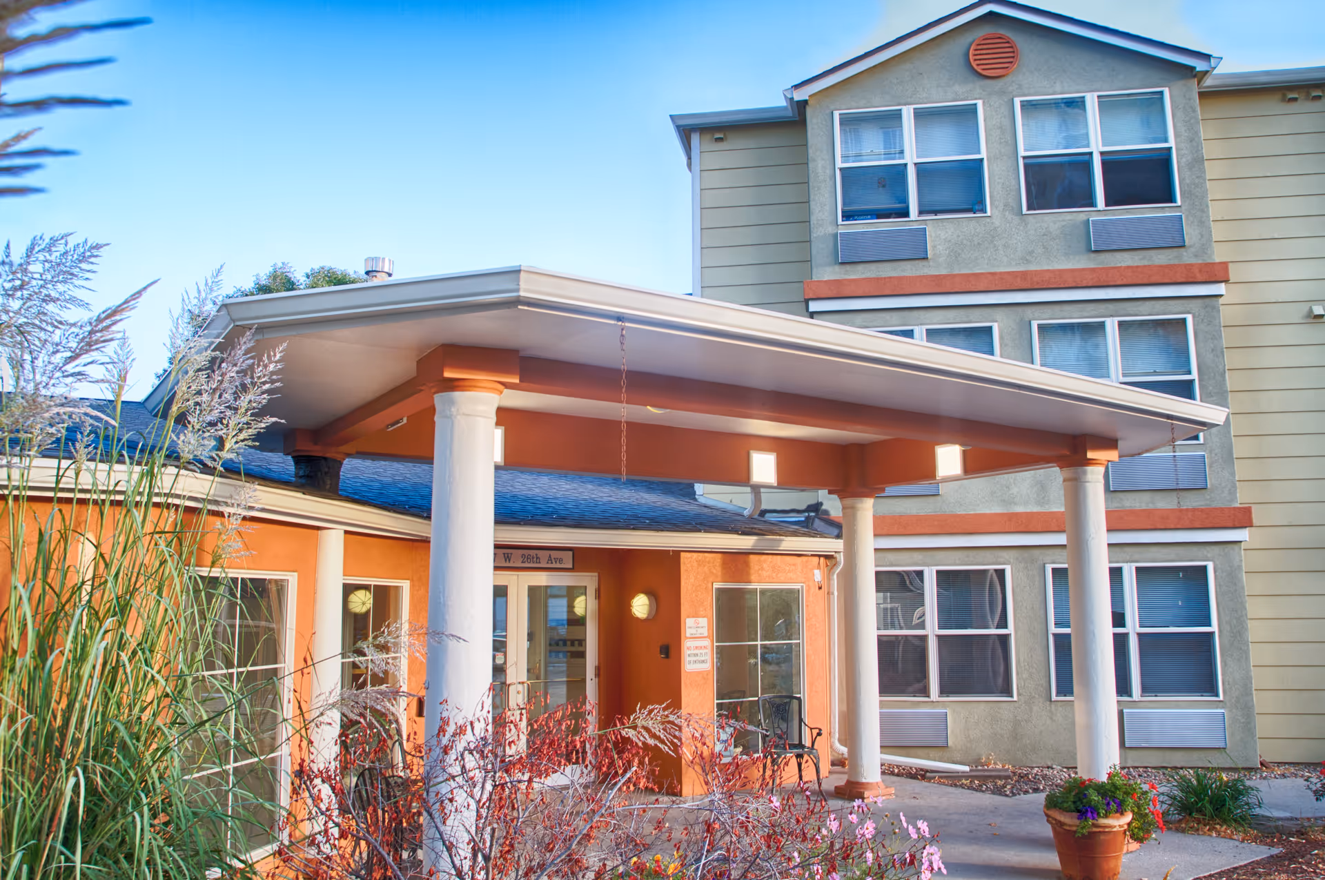 Front entrance of a multi-story senior living building with a covered portico supported by white columns, multiple windows, and landscaping.