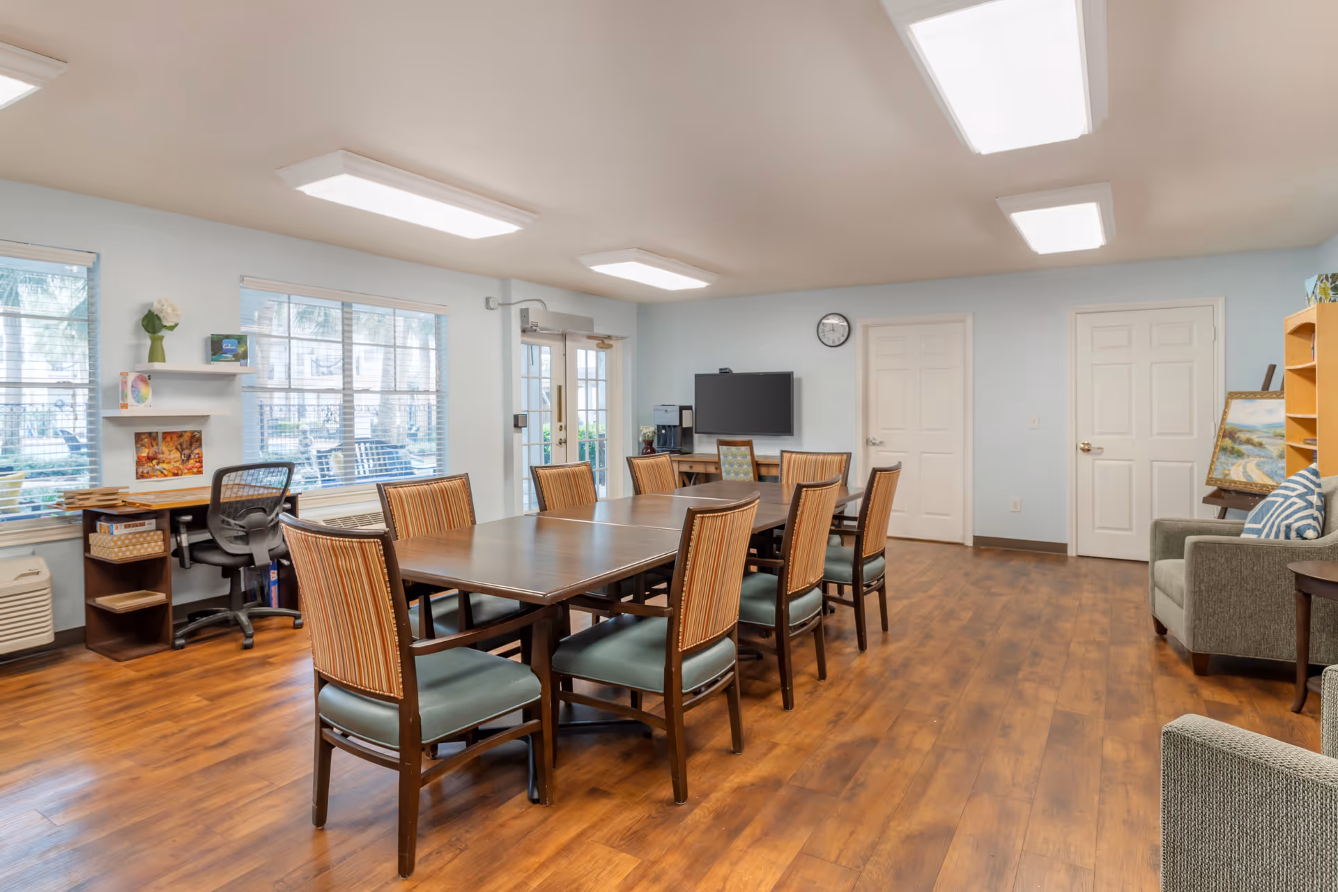 A well-lit community room with a large wooden table surrounded by eight chairs with striped backs and green seats. The room has light blue walls, wood flooring, and three white doors. There is a flat-screen TV mounted on the wall above a small desk with a chair, a coffee maker, and a clock. Windows with blinds allow natural light to enter, and there is a small seating area with a gray armchair and a painting on an easel.