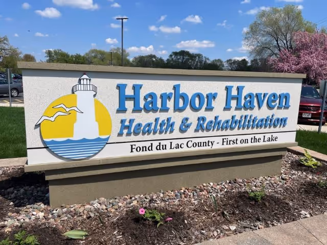 Outdoor sign for Harbor Haven Health & Rehabilitation with a lighthouse and seagulls logo, situated in a landscaped area with rocks and plants, under a blue sky with some clouds.