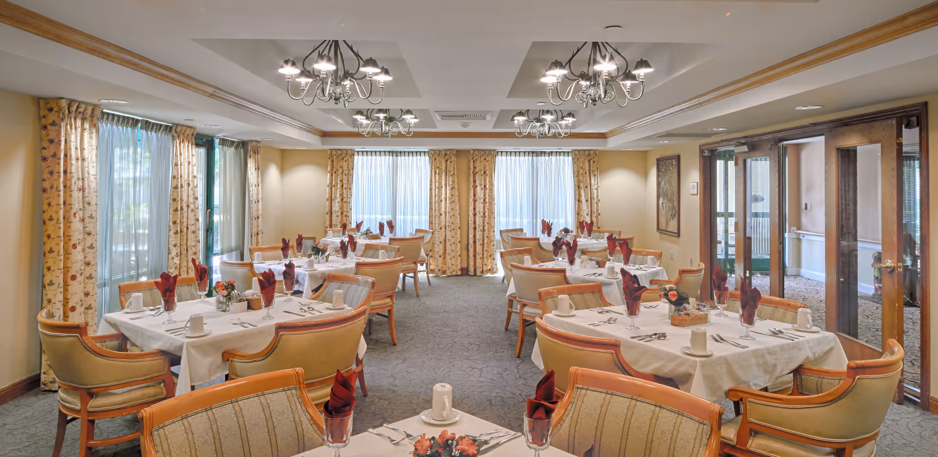 A well-lit dining room with several tables covered in white tablecloths, each set with folded red napkins, cups, and silverware. The room features beige upholstered chairs with wooden frames, floral curtains on large windows, and multiple chandeliers hanging from the ceiling. There is a glass door on the right side leading to another area.