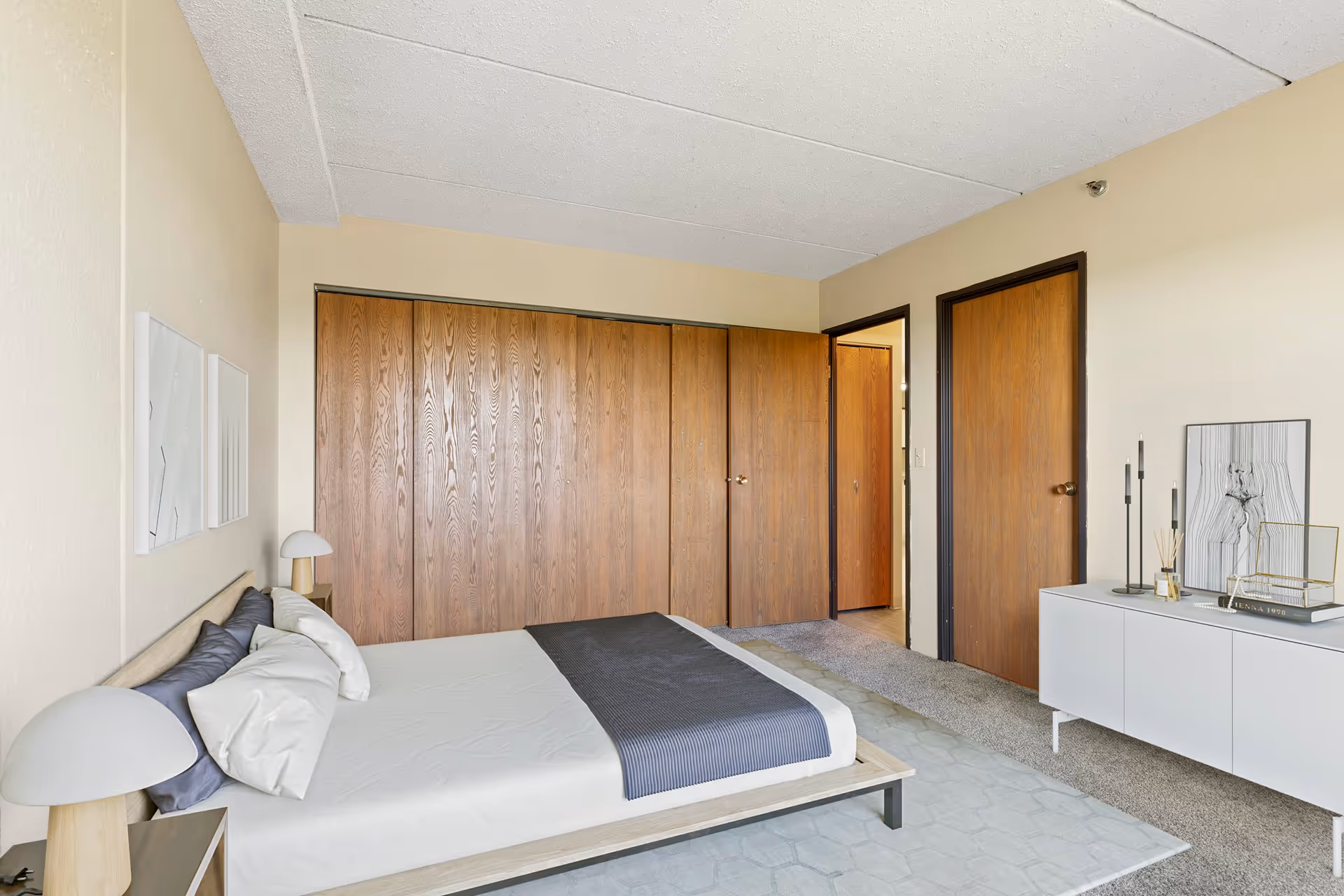 A bedroom with a bed featuring white and gray bedding, two bedside tables with modern lamps, a large wooden closet with multiple doors, a white cabinet with decorative items including candles and a framed artwork, and two wooden doors leading to other rooms.