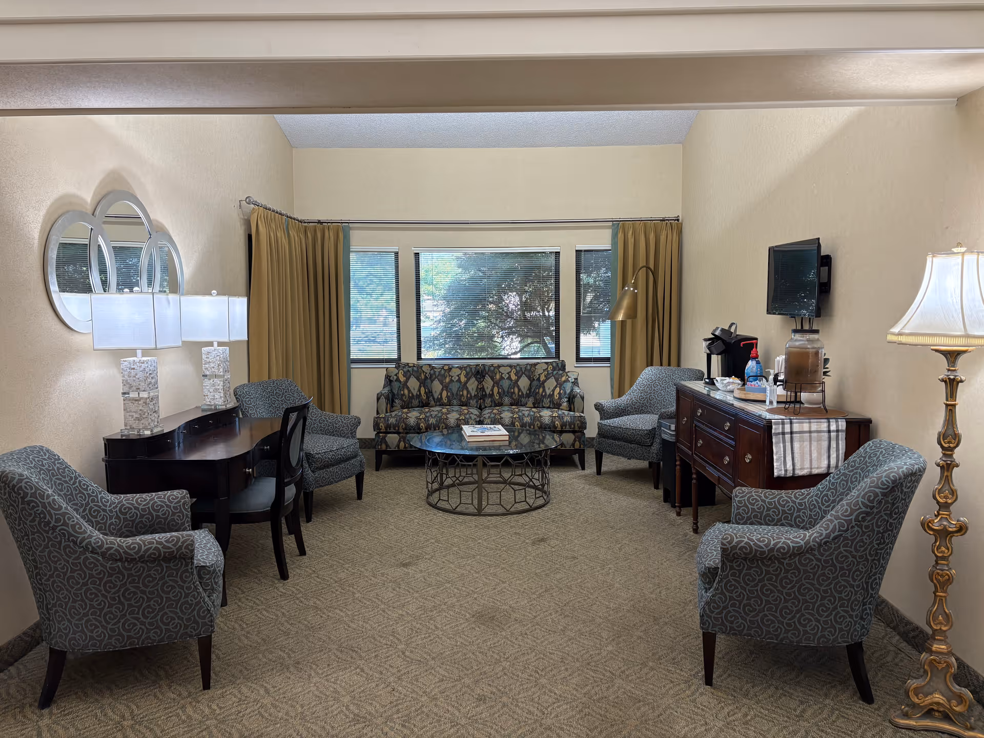 Well-lit communal living room with a patterned sofa and armchairs arranged around a coffee table, a sideboard with a coffee station, lamps, and a wall-mounted TV.