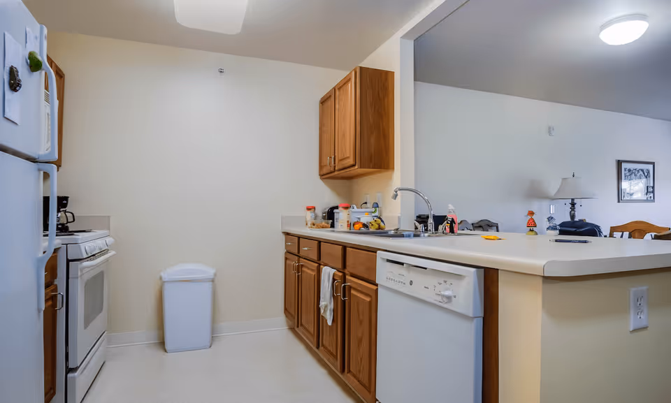 A kitchen area in a retirement village featuring wooden cabinets, a white refrigerator, a white stove, a white dishwasher, a trash bin, and a countertop with a sink and various items including a coffee maker and containers. The kitchen opens to a living area with a lamp and framed picture on the wall.