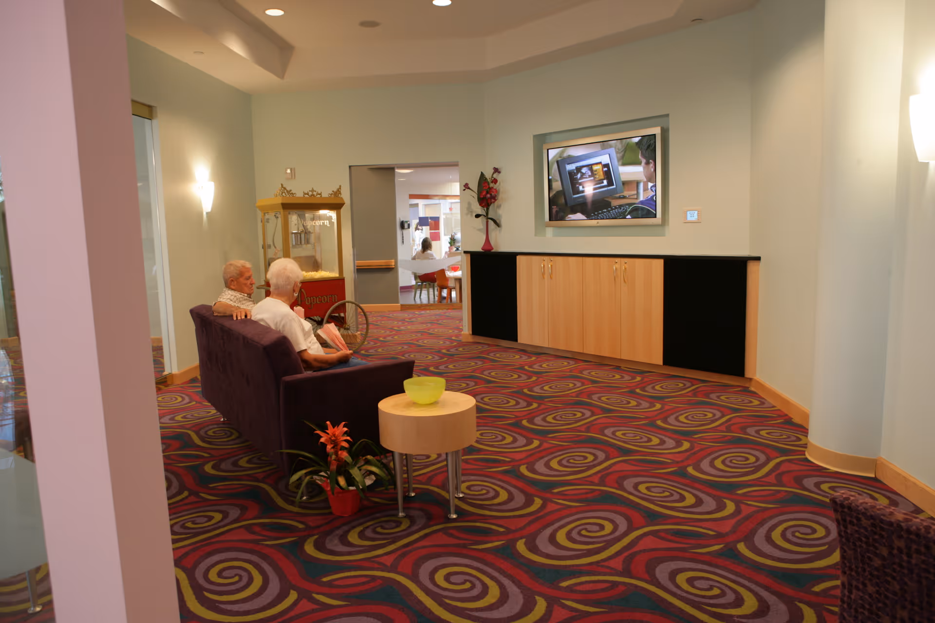 Two elderly people sit on a purple couch in a colorful carpeted lounge facing a wall-mounted TV and a popcorn machine.