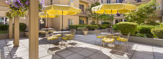 Sunny courtyard patio with yellow umbrellas, cushioned metal tables and chairs surrounded by shrubs and the assisted living building.