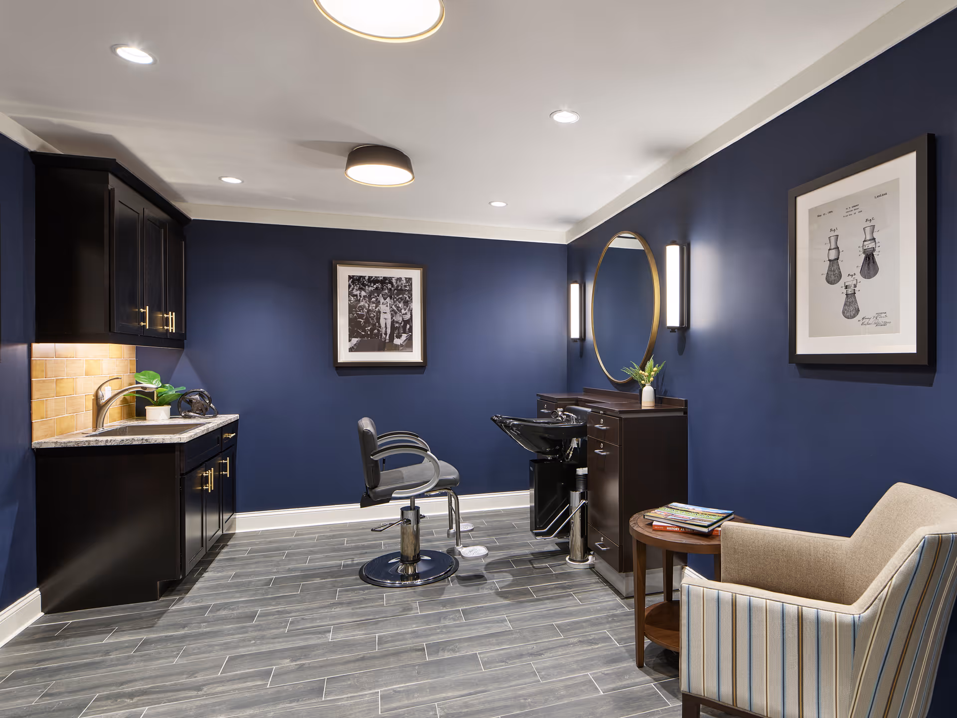 A modern salon area with dark blue walls, a styling chair in front of a black wash basin and a round mirror mounted on the wall. There is a beige armchair with striped upholstery next to a small round wooden table holding magazines. On the left side, there is a countertop with a sink, cabinets, and a small plant. The floor is covered with gray wood-like tiles, and the ceiling has recessed lighting and a round ceiling light.