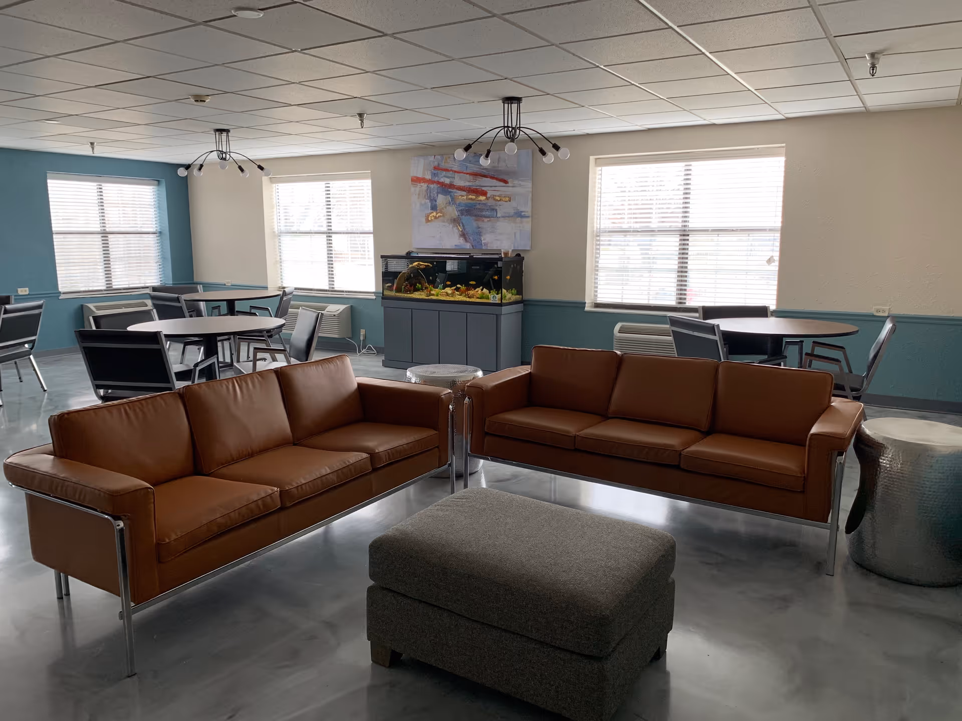 A common area with two brown leather sofas facing each other, a gray ottoman in the center, and several round tables with chairs near windows. There is an aquarium on a cabinet against the wall and a modern abstract painting above it. The room has a blue and white color scheme with large windows letting in natural light.