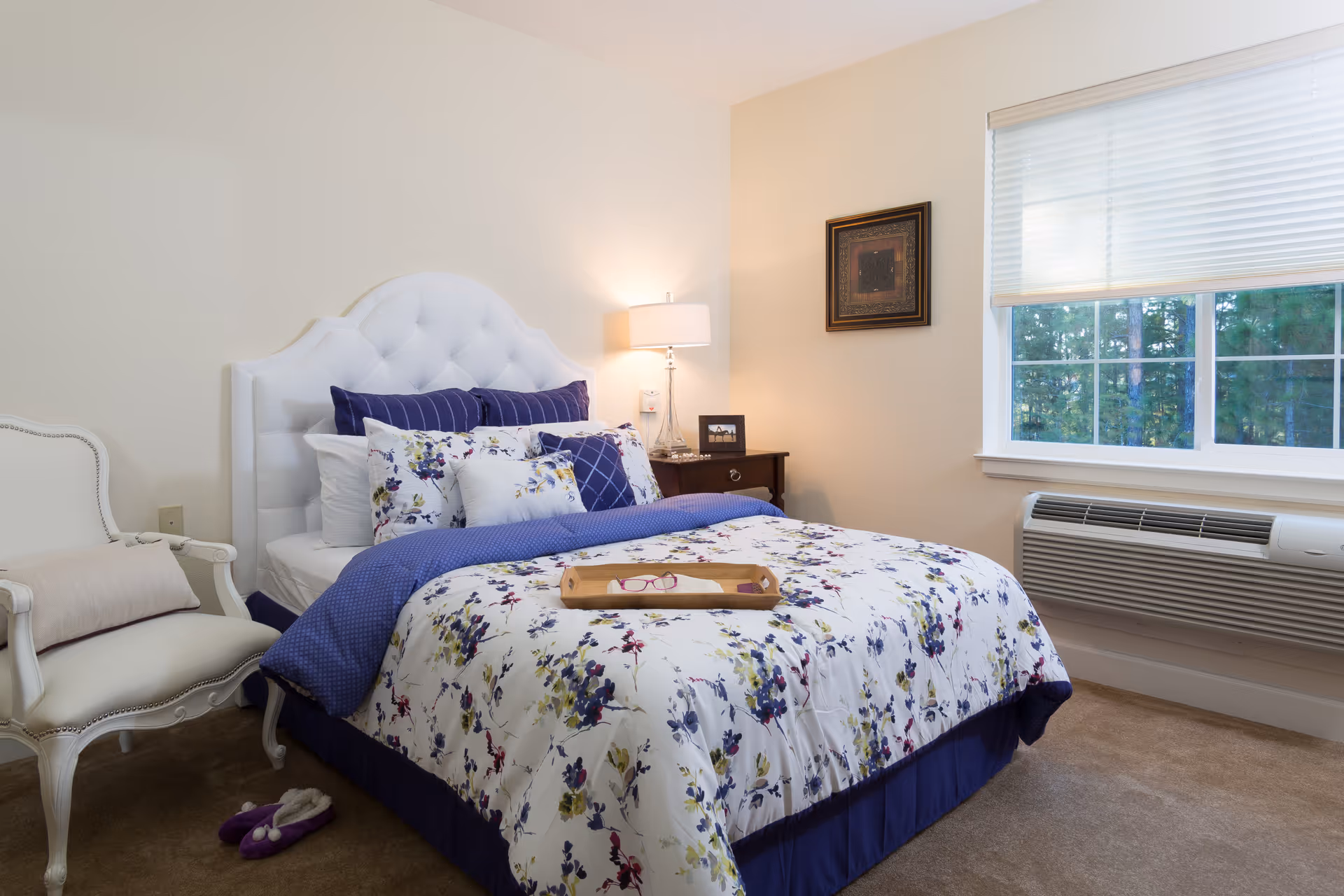 A cozy bedroom with a white tufted headboard bed dressed in floral and blue bedding. A wooden tray with glasses rests on the bed. To the left, there is a white armchair with a cushion and purple slippers on the carpeted floor. A nightstand with a lamp and a framed photo is next to the bed. A large window with white blinds lets in natural light, and an air conditioning unit is installed below the window.