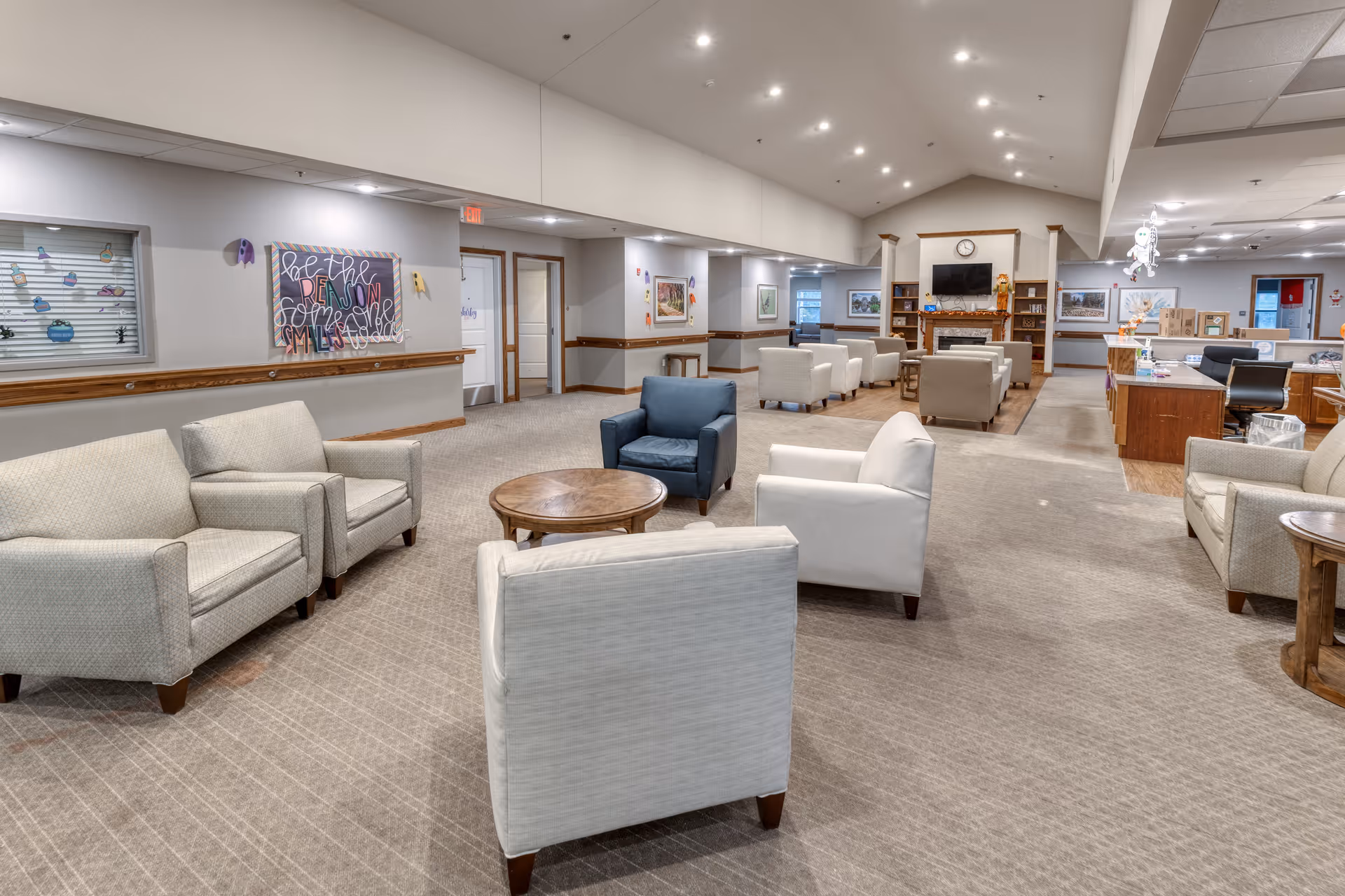 A spacious senior living common area with multiple armchairs arranged around small round wooden tables. The room features a carpeted floor, high ceiling with recessed lighting, and a fireplace with a TV mounted above it in the background. There are framed pictures on the walls and a reception or service desk area to the right.