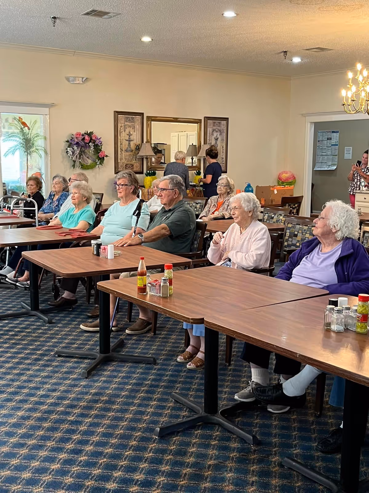 A group of elderly people sitting at tables in a common room with a carpeted floor and beige walls. Some are seated in chairs with patterned upholstery, and there are condiment bottles on the tables. In the background, two women are standing near a counter with a mirror and framed artwork on the wall. A chandelier is visible on the ceiling.
