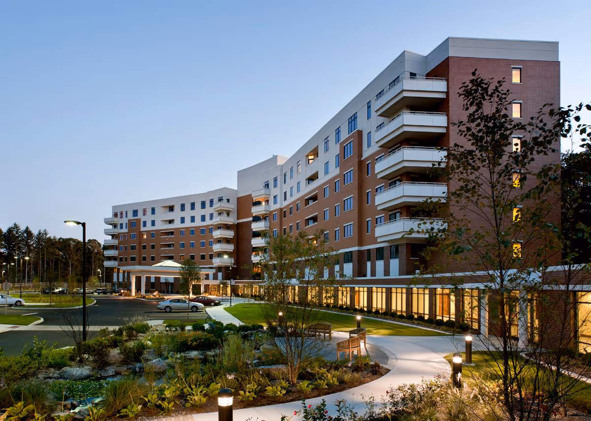 Exterior view of a large multi-story senior living facility building at dusk with landscaped garden, walking paths, benches, and parked cars in front.
