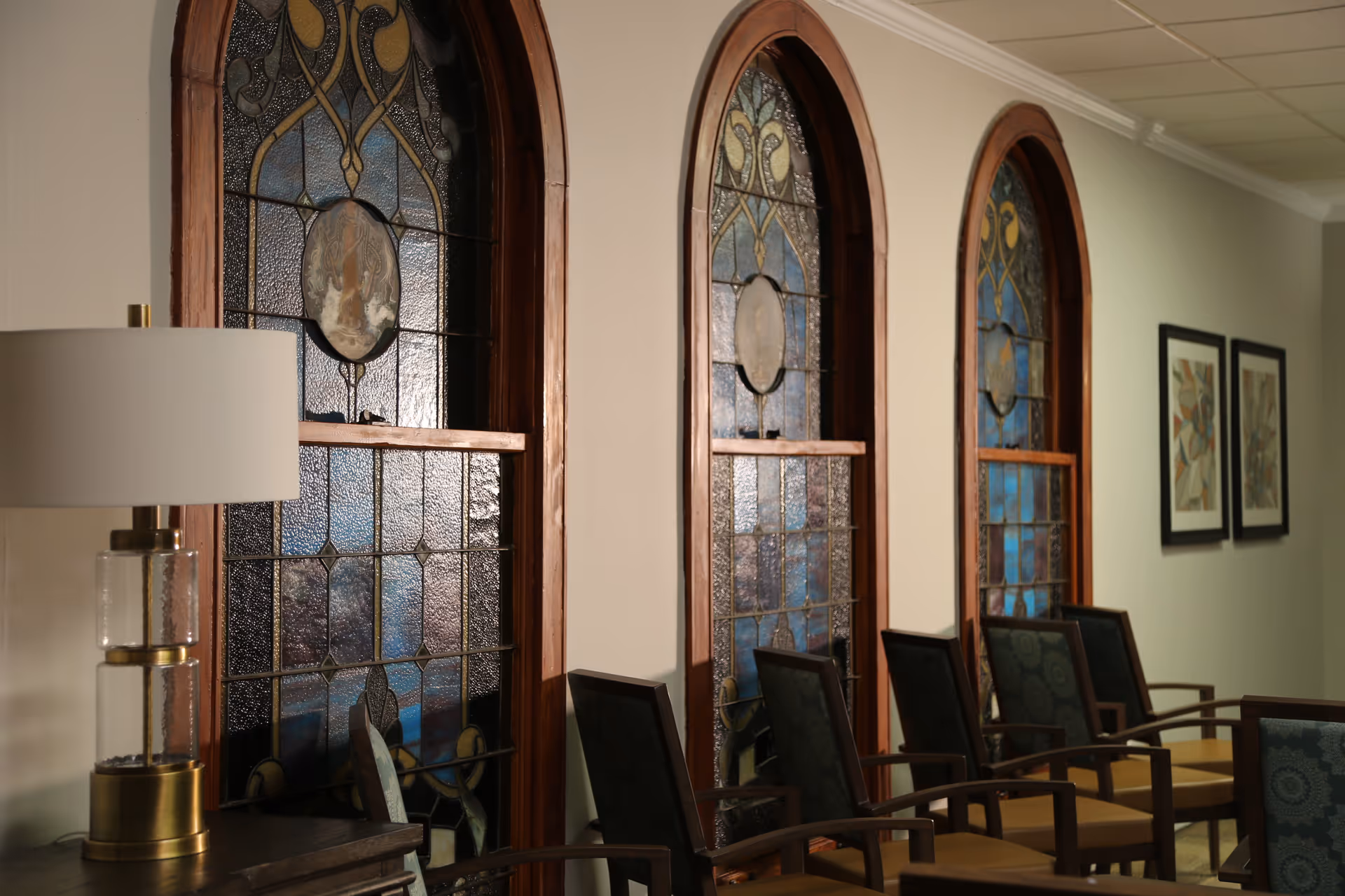 Interior view of a room with three tall arched stained glass windows along a wall. Below the windows are several wooden chairs with patterned cushions arranged in a row. To the left, there is a table with a modern lamp featuring a white shade and a gold and glass base. Two framed abstract artworks hang on the adjacent wall.