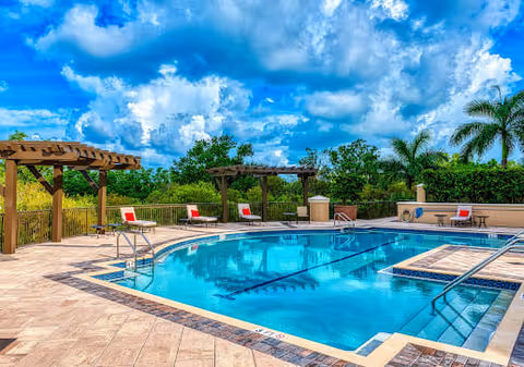 Outdoor swimming pool area with clear blue water, surrounded by a tiled deck. There are several lounge chairs with red cushions placed under wooden pergolas. Palm trees and green shrubbery are visible in the background under a partly cloudy blue sky.