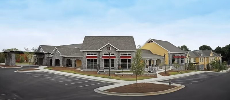 Exterior view of a senior living facility building with multiple connected sections, featuring stone and yellow siding, red awnings over windows, and a large parking lot in front under a partly cloudy sky.