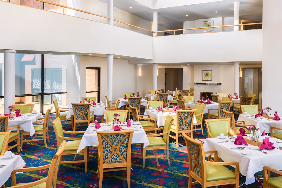 Bright and spacious dining room with multiple tables covered in white tablecloths, each set with pink folded napkins, plates, cups, and silverware. The room features wooden chairs with green cushions and decorative backs, colorful patterned carpet, large windows letting in natural light, and a two-story ceiling with white pillars and a balcony above.
