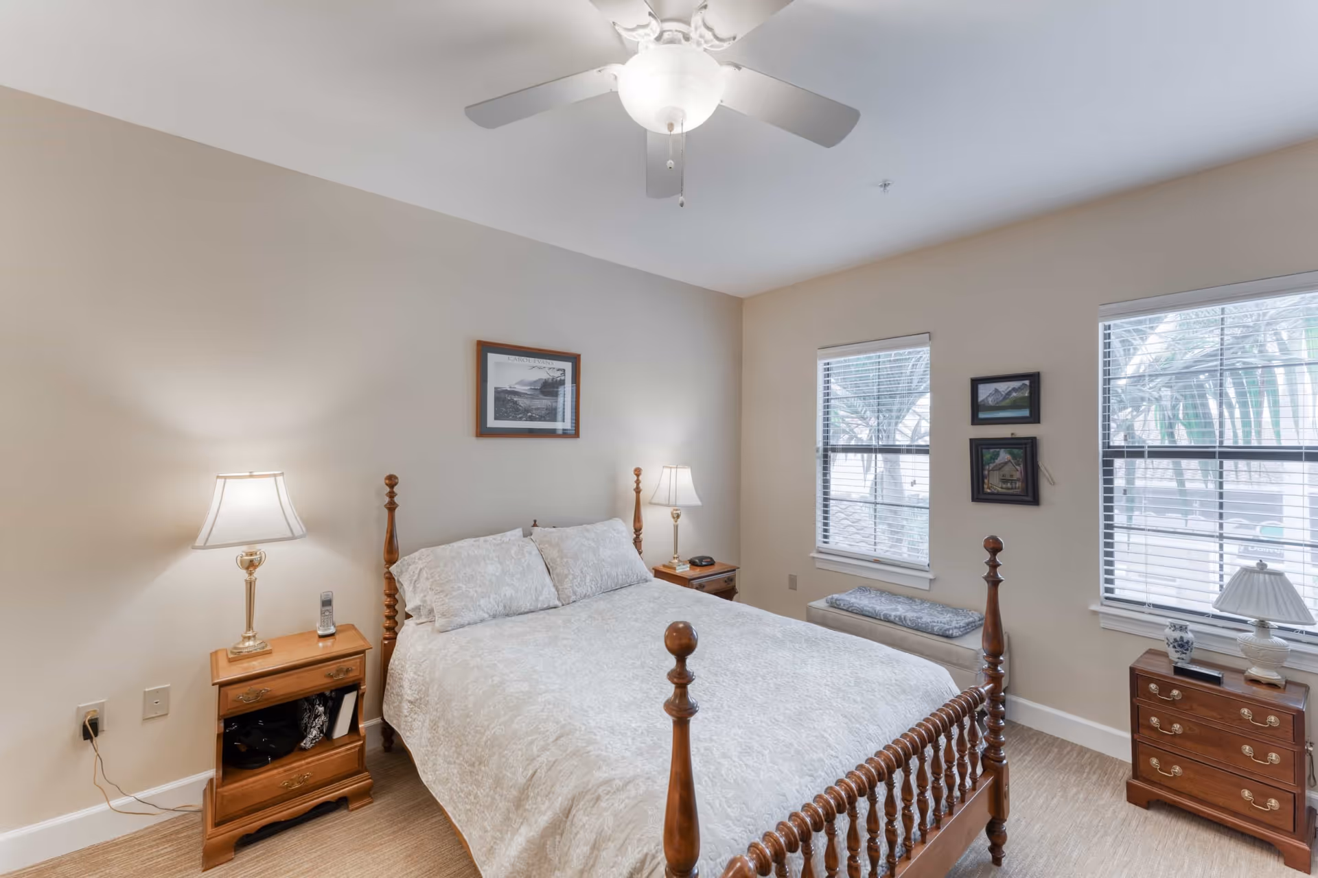 A cozy bedroom with a wooden four-poster bed covered in a white patterned bedspread. There are two wooden nightstands on either side of the bed, each with a lamp. The room has beige walls, two windows with white blinds, and a ceiling fan with a light fixture. There are framed pictures on the walls and a cushioned bench under one window.
