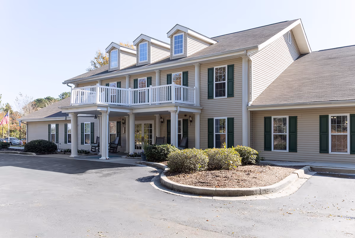 Exterior view of a two-story senior living facility building with beige siding, green window shutters, and a white balcony above the entrance. There are rocking chairs on the covered porch and some bushes in front of the building. The sky is clear and the parking area is visible in the foreground.