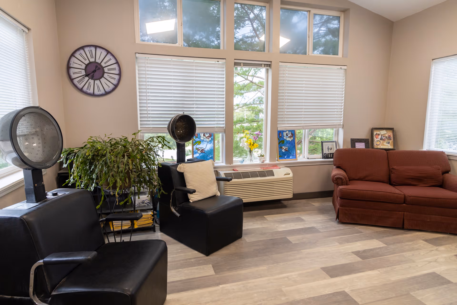 A cozy interior room with two black salon chairs equipped with hair dryers, a large green plant, a red couch with a pillow, a wall clock, and windows with white blinds partially open showing some greenery outside.