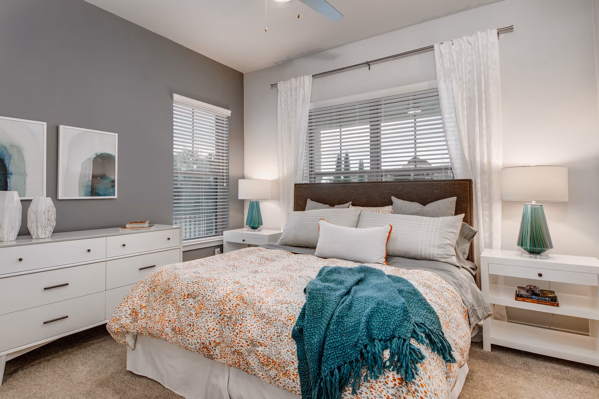 A cozy bedroom with a large bed featuring multiple pillows and a patterned comforter with a teal throw blanket. The room has two windows with white blinds and white curtains. There are two white nightstands on either side of the bed, each with a teal lamp. A white dresser with decorative vases and framed abstract art is positioned against a gray accent wall.
