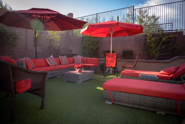 Outdoor patio area with red cushioned wicker lounge chairs, sofas, and umbrellas on artificial grass, surrounded by a high fence and some greenery.